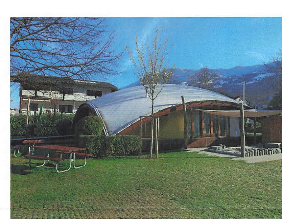 A curved, silver-roofed building on a lawn with mountains behind it.